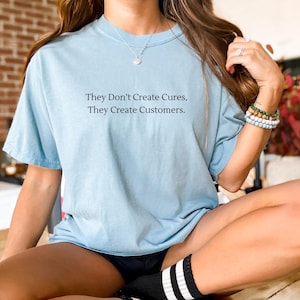 May include: A light blue t-shirt with the text "They Don't Create Cures, They Create Customers." printed on the front.