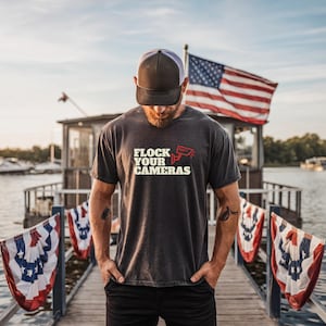 May include: A charcoal gray short-sleeve t-shirt featuring the text "FLOCK YOUR CAMERAS" in white lettering with a red security camera icon. The shirt is paired with a two-tone gray and white baseball cap and dark pants.