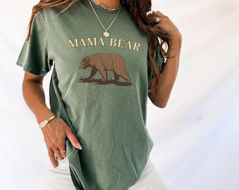May include: Green t-shirt with the text "MAMA BEAR" in a serif font above a brown bear graphic. The bear is walking on a brown patch of grass. The shirt has short sleeves and a relaxed fit.