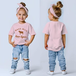 Puede incluir: Una camiseta rosa con un gr&aacute;fico de vaca marr&oacute;n y el texto "Make Milk Raw Again". La camiseta la lleva puesta un ni&ntilde;o con una diadema rosa y unos vaqueros azules.