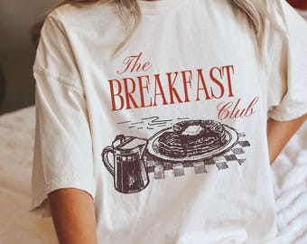 May include: Cream-colored t-shirt featuring the text "The Breakfast Club" in red script. Below the text is a vintage-style illustration of a stack of pancakes with butter, a coffee pot, and a checkered tablecloth. The shirt has short sleeves.