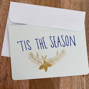 May include: A white greeting card with a matching envelope. The card has the phrase "'TIS THE SEASON" in blue script, with a gold-colored sprig and holly design below the text. The card is on a wooden surface.