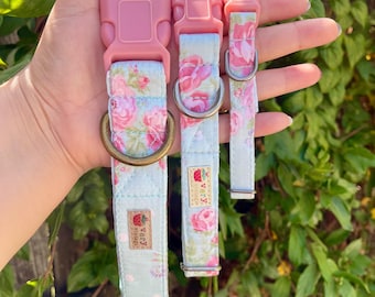 May include: Three dog collars in varying sizes. The collars feature a light blue floral pattern with pink roses and green leaves. Each collar has a pink plastic buckle and a metal D-ring. The collars are held up against a background of green foliage.