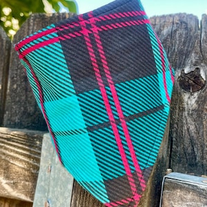 May include: A turquoise, black, and pink plaid dog bandana. The bandana is folded and appears to be made of fabric. The plaid pattern consists of turquoise and pink lines over a black background. The bandana is hanging on a wooden fence.