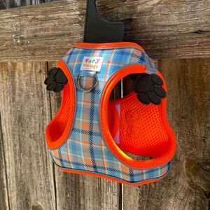 Puede incluir: Arn&eacute;s para perro de cuadros naranja y azul con un dise&ntilde;o de huella de pata negra y las palabras "very vintage" en la parte delantera. El arn&eacute;s est&aacute; colgado de un gancho negro.