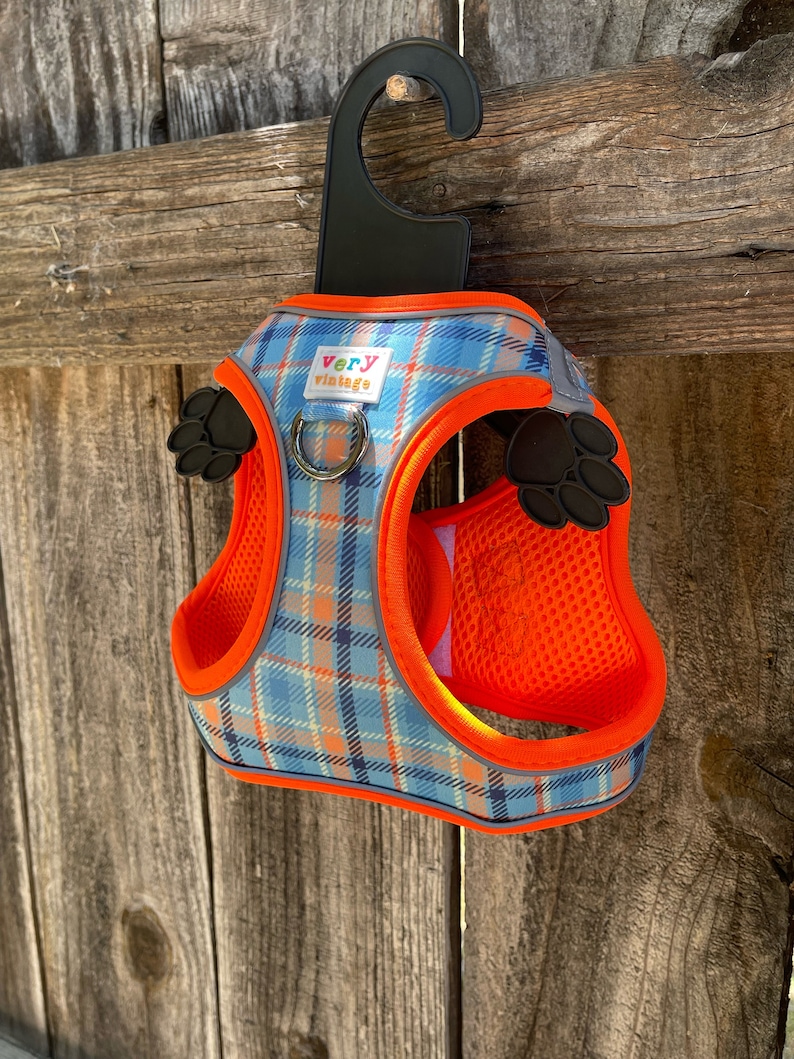 Puede incluir: Arn&eacute;s para perro de cuadros naranja y azul con un dise&ntilde;o de huella de pata negra y las palabras "very vintage" en la parte delantera. El arn&eacute;s est&aacute; colgado de un gancho negro.