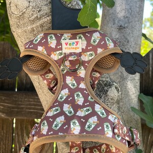 May include: A brown dog harness with a pattern of white ice cream cups and green leaves. The harness has a brown strap with a metal buckle and a brown mesh panel. The harness has a small tag that says "very vintage".