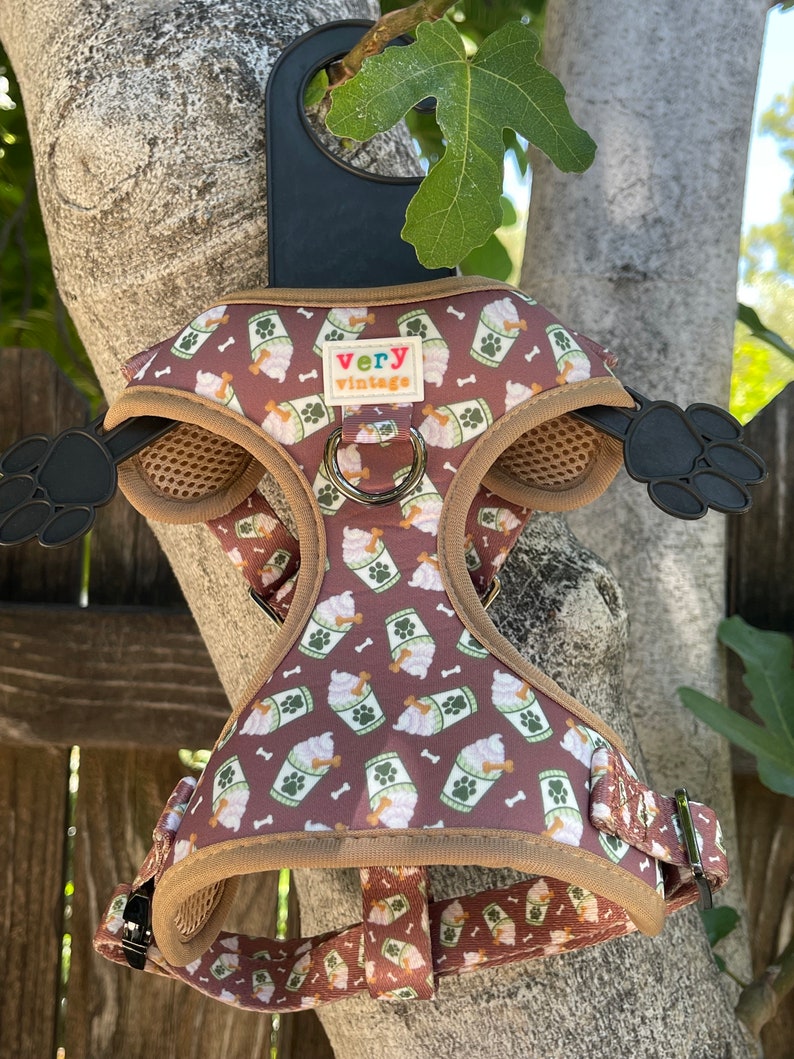 May include: A brown dog harness with a pattern of white ice cream cups and green leaves. The harness has a brown strap with a metal buckle and a brown mesh panel. The harness has a small tag that says "very vintage".