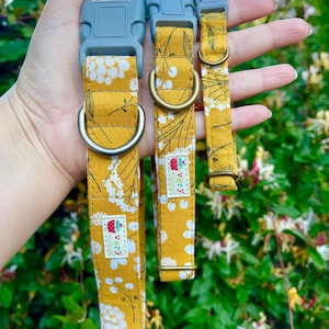 Three handmade pet collars featuring a mustard yellow background with white and gray floral accents, chocolate brown buckles, and antique brass and antique silver hardware. Displayed in a hand against a lush green garden background with soft sunlight.