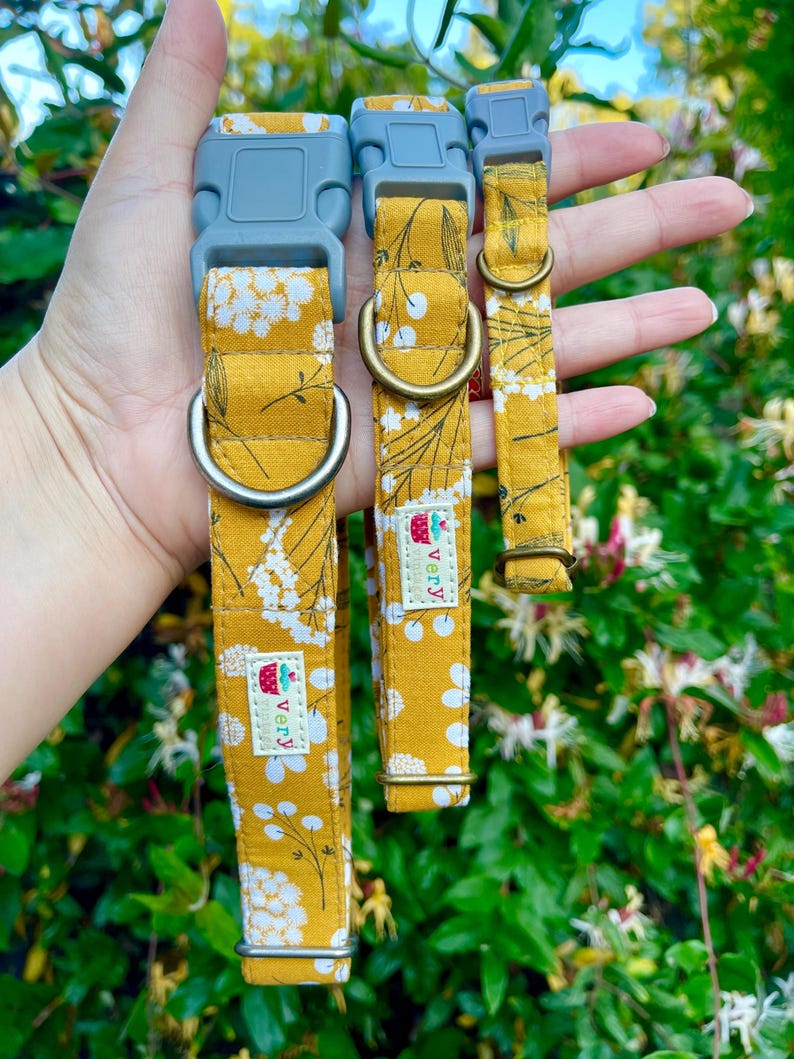 Three handmade pet collars featuring a mustard yellow background with white and gray floral accents, chocolate brown buckles, and antique brass and antique silver hardware. Displayed in a hand against a lush green garden background with soft sunlight.