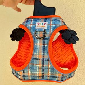 Puede incluir: Arn&eacute;s para perro de cuadros naranja y azul con una huella de pata negra a cada lado. El arn&eacute;s tiene un anillo en D de metal negro y un gancho de pl&aacute;stico negro. El arn&eacute;s tiene el texto "very vintage" en la parte delantera.