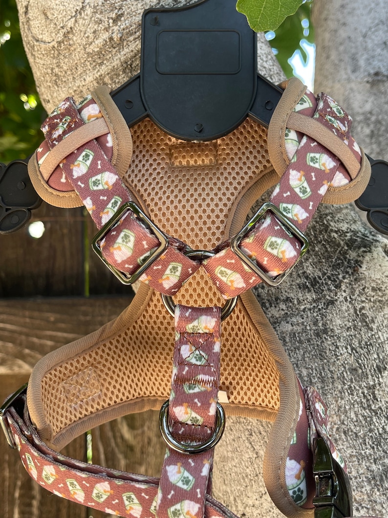 May include: A brown dog harness with a white and green pattern of ice cream cones and bones. The harness has a mesh back panel and a metal buckle.