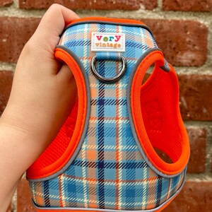 Plaid dog harness in orange, blue, and gray, held in hand in front of red brick wall