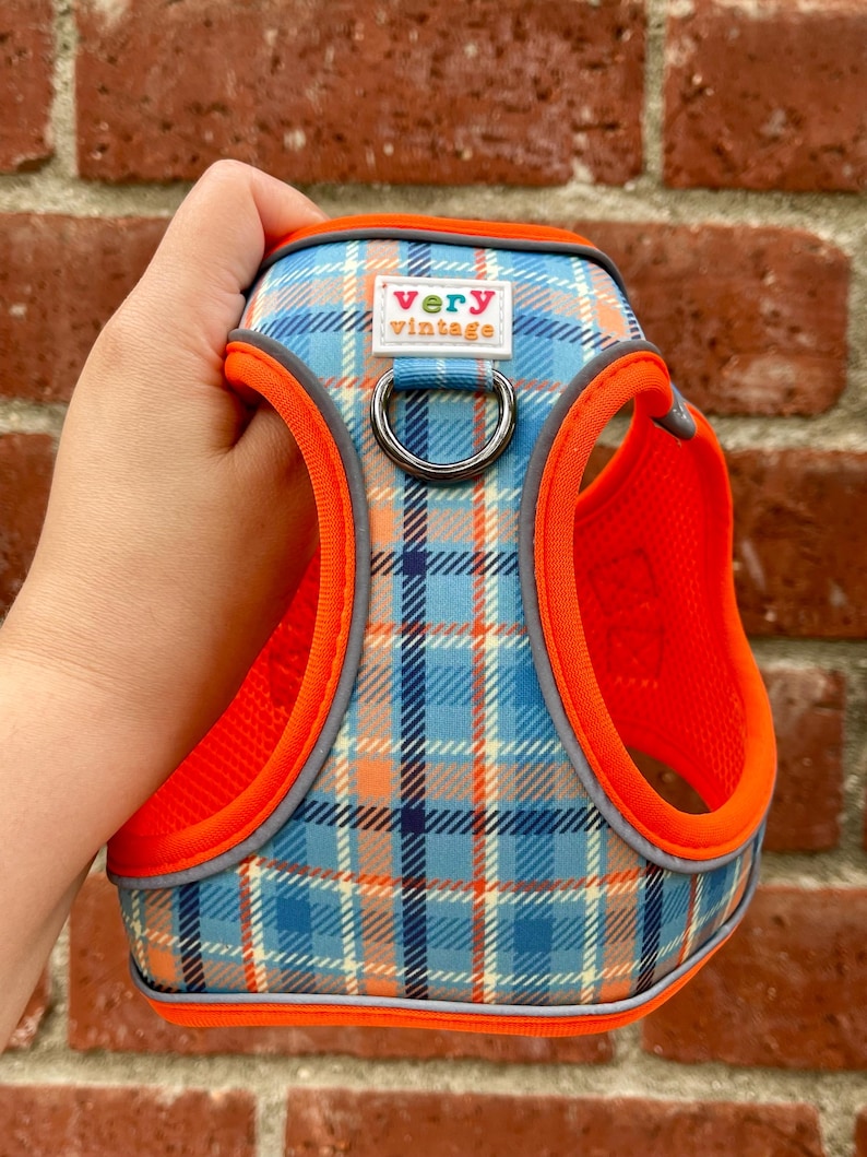 Plaid dog harness in orange, blue, and gray, held in hand in front of red brick wall