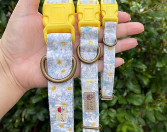 May include: Three dog collars with yellow buckles and a white floral pattern. The collars are lined up in order of size, with the largest collar on the left and the smallest collar on the right. The collars have a "Very" label with a strawberry graphic.
