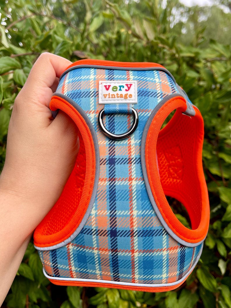 Blue and orange plaid dog harness with orange mesh lining, shown in hand against leafy green background