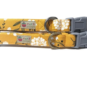 Two pet collars with mustard yellow background and white wildflower print, featuring gray buckles and antique brass and antique silver hardware. Displayed on a white background.