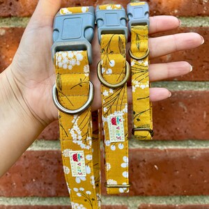 Three handmade pet collars with a mustard yellow base and delicate white and gray floral pattern, chocolate brown buckles, and antique brass and antique silver hardware. Displayed in a hand in front of a weathered red brick wall. Handmade in Los Angeles.