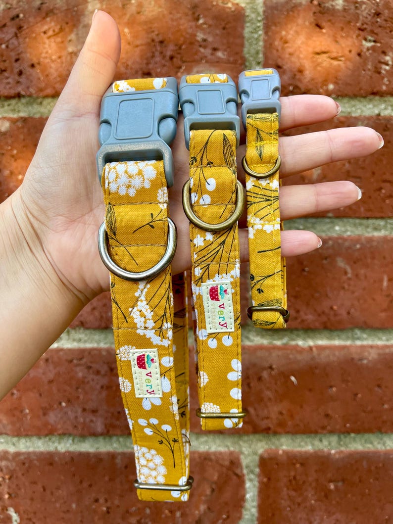 Three handmade pet collars with a mustard yellow base and delicate white and gray floral pattern, chocolate brown buckles, and antique brass and antique silver hardware. Displayed in a hand in front of a weathered red brick wall. Handmade in Los Angeles.