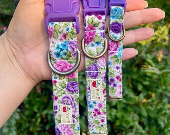 May include: Three purple dog collars with a floral pattern and a purple buckle. The collars have a white background with pink, purple, and blue flowers. The collars have a small label that says "very" with a strawberry graphic.