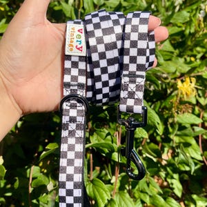 May include: A black and white chequered dog lead featuring a rectangular white fabric tag with the text very vintage in colourful letters. The lead includes a silver metal D-ring and a black metal swivel snap hook attached to the woven fabric strap.