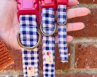 May include: Three dog collars in different sizes, all with a blue and white gingham pattern and red buckles. The collars have brass D-rings and a small label with the text "very" on them.