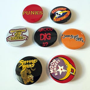 Könnte beinhalten: Sieben runde, bunte Anstecknadeln mit verschiedenen Bandnamen und Logos. Die Knöpfe zeigen die Namen "Punks", "Electric Eliminators", "Ban Cortianbt Rangers", "Can You Dig It?", "Jones Street Boys", "Savage Shuns" und "Moon Runners".