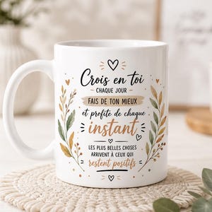 Peut inclure: Un mug en céramique blanc orné du texte "Crois en toi chaque jour, fais de ton mieux et profite de chaque instant, les plus belles choses arrivent à ceux qui restent positifs" entouré d'illustrations botaniques de feuilles décoratives.