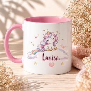 Puede incluir: Taza de cerámica blanca con interior y asa rosa. La taza presenta una colorida ilustración de un unicornio sentado en una nube con un arcoíris, corazones y estrellas. El nombre "Louisa" está escrito en cursiva.