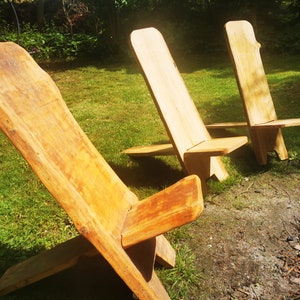Op de afbeelding: Drie houten stoelen met een rustieke, natuurlijke afwerking zijn in een rij op een gazon geplaatst. De stoelen zijn gemaakt van één stuk hout en hebben een eenvoudig, minimalistisch ontwerp.