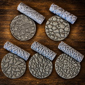 Puede incluir: Un conjunto de cinco rodillos cilíndricos grises y cinco discos circulares a juego, cada uno con texturas en relieve distintas que incluyen patrones de vetas orgánicas, adoquines, fragmentos geométricos y superficies de piedra agrietada.