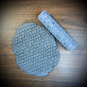May include: A cylindrical rolling pin with a textured grid pattern and a flat, circular piece of gray clay featuring an identical embossed cobblestone design. The tool and the clay impression both display a repeating square tile motif.