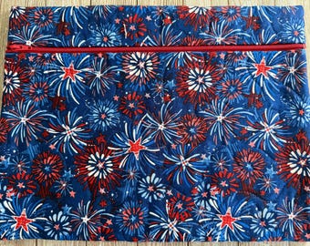 May include: A rectangular quilted fabric pouch featuring a dark blue background with a repeating pattern of red, white, and blue fireworks and stars. The pouch includes a horizontal red zipper closure extending across the top section.