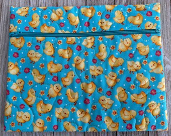 May include: A quilted, rectangular pouch with a teal zipper. The pouch features a repeating pattern of yellow chicks, pink flowers, and white polka dots on a teal background. The stitching creates a quilted effect.