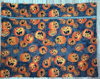 May include: A quilted fabric pouch with a zipper closure, featuring a repeating pattern of orange jack-o'-lanterns on a dark blue background. The pumpkins have various carved faces. The pouch is rectangular and suitable for storage.