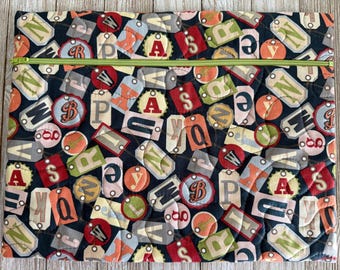 May include: A rectangular quilted fabric pouch featuring a dark navy blue background covered in a repeating pattern of colorful, multi-shaped tags, each printed with various letters of the alphabet. A bright lime green zipper runs across the top.