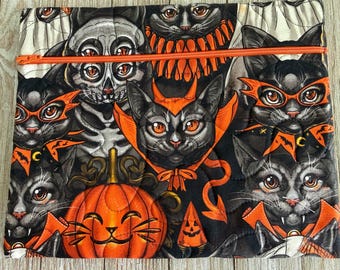 May include: A quilted zippered pouch featuring a repeating pattern of black cats dressed in orange Halloween costumes, including vampire capes and masks, interspersed with orange jack-o'-lantern pumpkins on a black background with orange stitching.