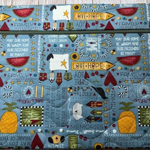 May include: A rectangular quilted fabric pouch with a zipper closure, featuring a light blue background printed with yellow pineapples, red watermelons, white sheep, small houses, and text including "LOVE HOPE," "FAMILY," and "SIMPLE JOY."
