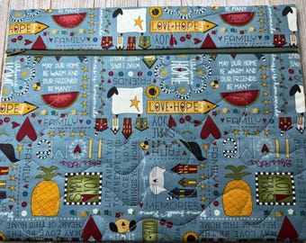 May include: A rectangular quilted fabric pouch with a zipper closure, featuring a light blue background printed with yellow pineapples, red watermelons, white sheep, small houses, and text including "LOVE HOPE," "FAMILY," and "SIMPLE JOY."