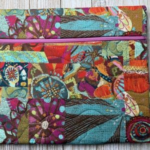 May include: A rectangular quilted fabric pouch featuring a vibrant, multi-colored abstract pattern with shades of teal, magenta, orange, and brown. The pouch includes a horizontal pink zipper closure across the top section of the quilted textile.