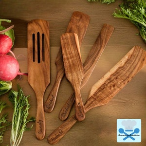 May include: Set of six wooden kitchen spatulas with personalised names, including "Sandy", "Rachel", "Hubert", "Emma", and two more "Rachel" names. The spatulas are made of natural wood with a brown colour and a grain pattern.