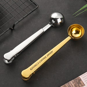 May include: Two coffee scoops with clips. One is silver and the other is gold. The gold scoop has the text "Rafael's Coffee Spoon" engraved on it.