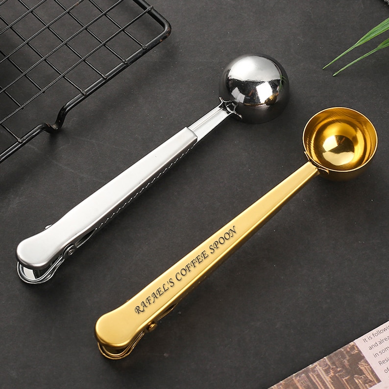 May include: Two coffee scoops with clips. One is silver and the other is gold. The gold scoop has the text "Rafael's Coffee Spoon" engraved on it.