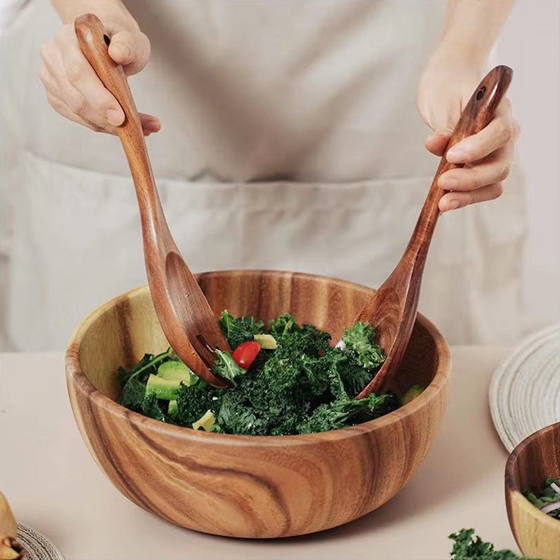 May include: A wooden salad bowl with a green salad, red tomatoes, and avocado inside. Two wooden salad servers are being used to toss the salad.