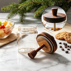 May include: A clear glass honey jar with a dark wood lid and an attached wooden honey dipper. The lid features a circular knob and the engraved text "Life is the honey". The jar is empty, while a circular inset shows the lid closed on a jar.