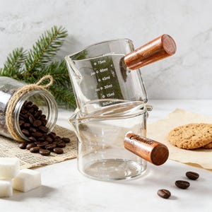 Könnte beinhalten: Zwei transparente Glasbecher mit Holzgriffen, übereinander gestapelt. Jeder Becher hat Messmarkierungen in Millilitern. Kaffeebohnen, Zuckerwürfel und Kekse sind ebenfalls im Bild, was auf ein Kaffee-Thema hindeutet.