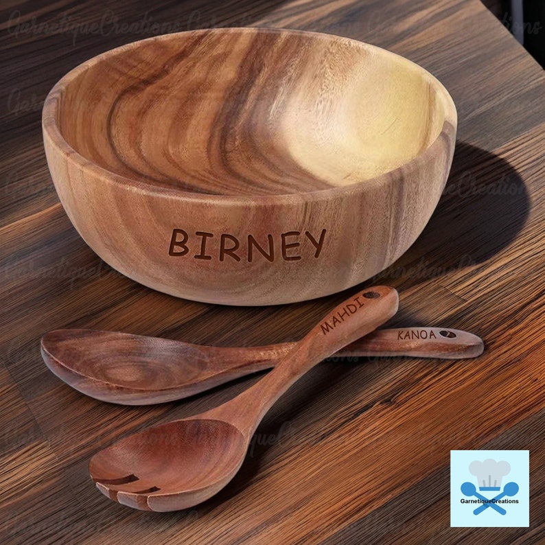 May include: A wooden salad bowl with the name "BIRNEY" engraved on the side, accompanied by two wooden spoons with the names "MAHDIA" and "KANOA" engraved on the handles. The bowl and spoons are on a wooden surface.