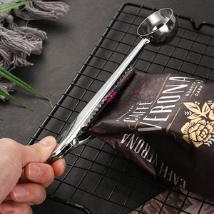 May include: A silver metal coffee scoop and bag clip with a serrated edge. The clip is holding a bag of coffee beans labeled "CAFFE VERONA" with the text "ROASTY & DARK".