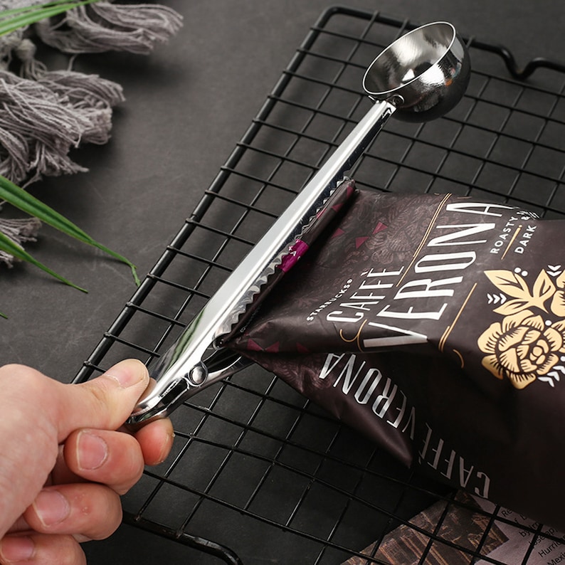 May include: A silver metal coffee scoop and bag clip with a serrated edge. The clip is holding a bag of coffee beans labeled "CAFFE VERONA" with the text "ROASTY & DARK".
