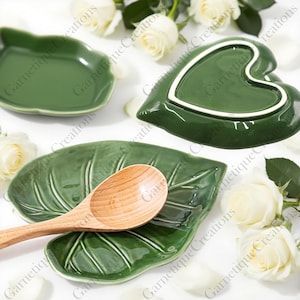 Könnte beinhalten: Grüne Keramikschalen in Blattform und eine herzförmige Schale mit einem Holzlöffel. Die Schalen sind glänzend grün. Weiße Rosen und Blütenblätter sind um die Schalen herum verstreut.